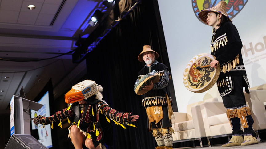 An indoor image of Spakwus Slolem (translated as ”Eagle Song Dancers”) from the Squamish Nation wearing traditional regalia and performing a song, with drumming, singing and dancing.
