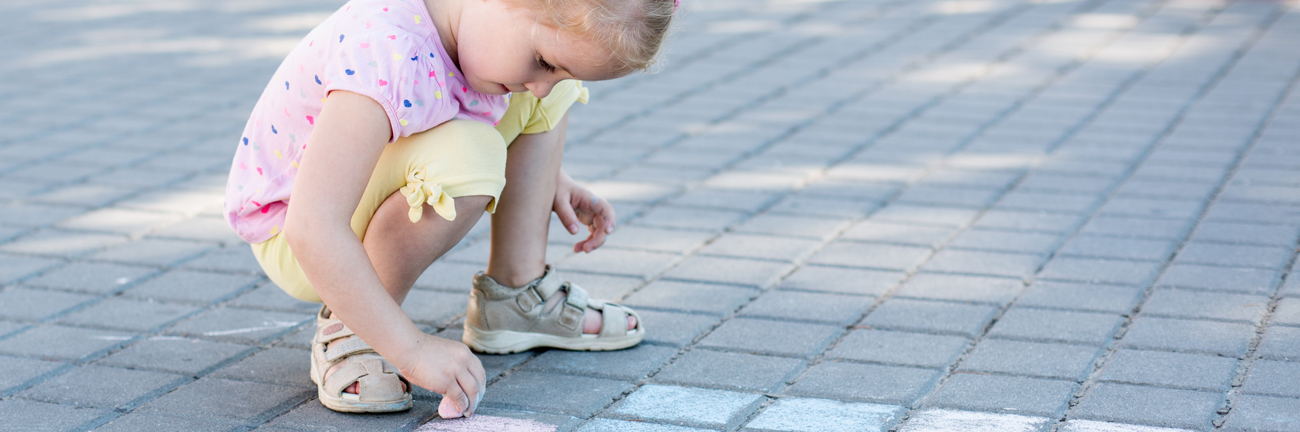 An outdoor image of a child's feet standing on a brick pathway, with their hand colouring on the ground using chalk