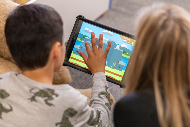 A child plays Dino Island on a tablet while an adult looks on.