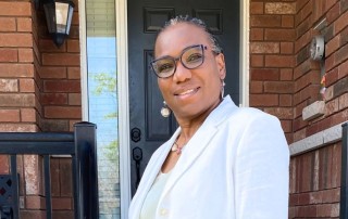 Sheila, a black woman wearing a white blazer, looks on, smiling.