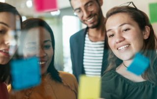 A group of four individuals collaborates enthusiastically, standing in front of a glass wall covered with colourful sticky notes. They are smiling and engaged, appearing to brainstorm or share ideas. The setting looks like a modern workspace, with bright lighting and a casual atmosphere.