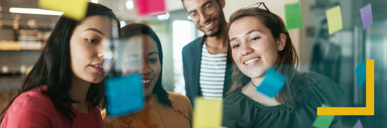 A group of four individuals collaborates enthusiastically, standing in front of a glass wall covered with colourful sticky notes. They are smiling and engaged, appearing to brainstorm or share ideas. The setting looks like a modern workspace, with bright lighting and a casual atmosphere.