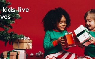 Two children in festive pajamas exchange gifts by a Christmas tree with stacked presents. Warm, joyful atmosphere. Text reads "Helping all kids live their best lives."