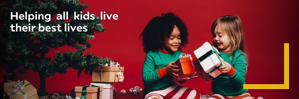 Two children in festive pajamas exchange gifts by a Christmas tree with stacked presents. Warm, joyful atmosphere. Text reads "Helping all kids live their best lives."