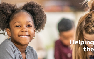 Smiling young girl with curly hair in a classroom setting, surrounded by other children. Text reads, "Helping all kids live their best lives."