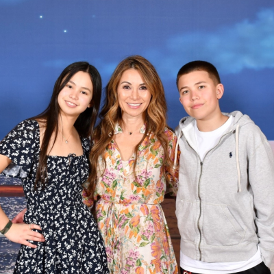 Sarah Jane Lee is in a floral dress and stands between a girl in a dark patterned dress and a boy in a gray hoodie. They smile against a backdrop of an ocean scene.