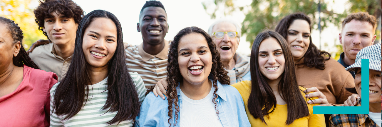 A diverse group of people standing together outdoors, smiling and looking at the camera. The mood is joyful and inclusive, with trees in the background.